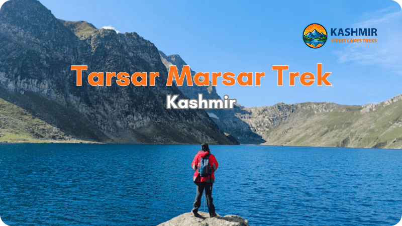 Trekker in red jacket standing on rocks beside pristine Tarsar Lake with blue waters and mountain peaks in background